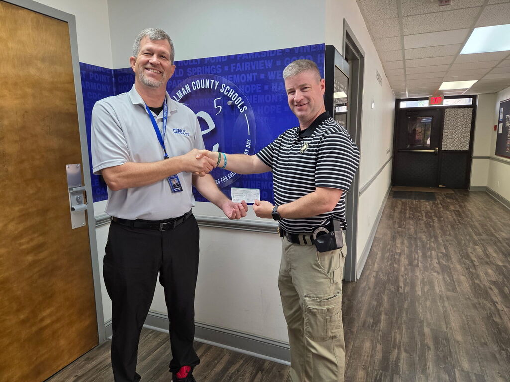 Two men in a hallway, shaking hands. One holds a certificate. Behind them, a blue sign and a wooden door.