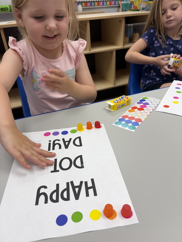 Two young girls, one in pink, another in blue, work at a table. The table displays a board with colored words and gummy pieces.