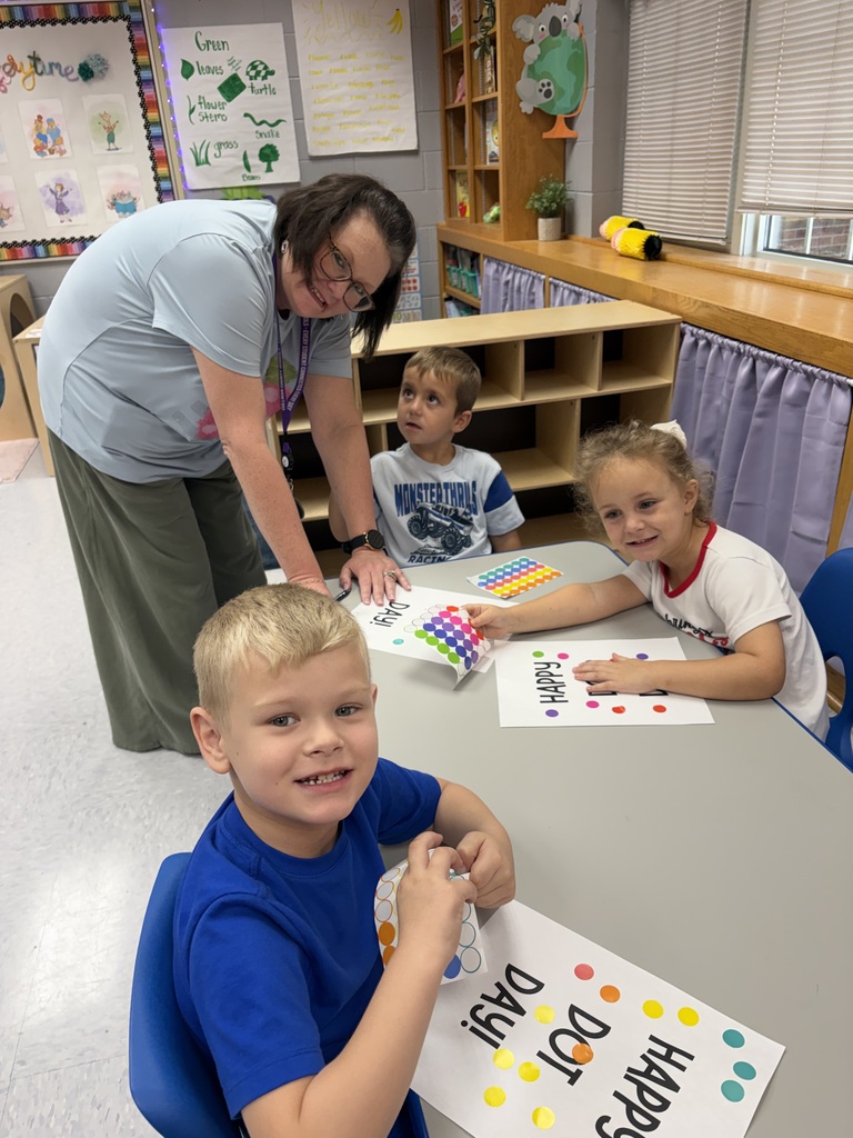 An adult helps two children, one holding a sign that says "Happy", work on a craft project at a table.