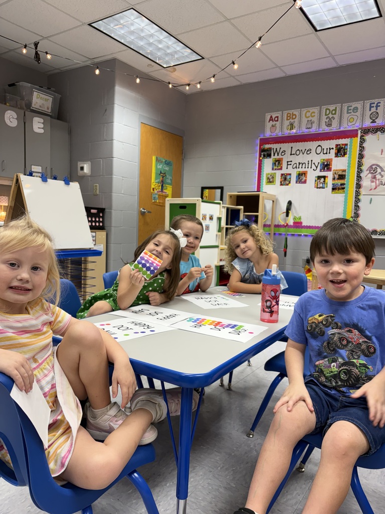 Six children sit at a table in a classroom. One holds a string of beads.