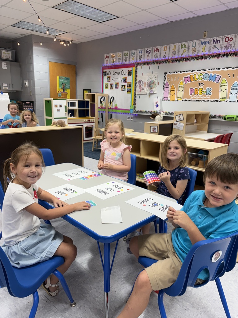 Four children sit at a table in a classroom, each holding papers and smiling. Behind them, a whiteboard is on the wall.