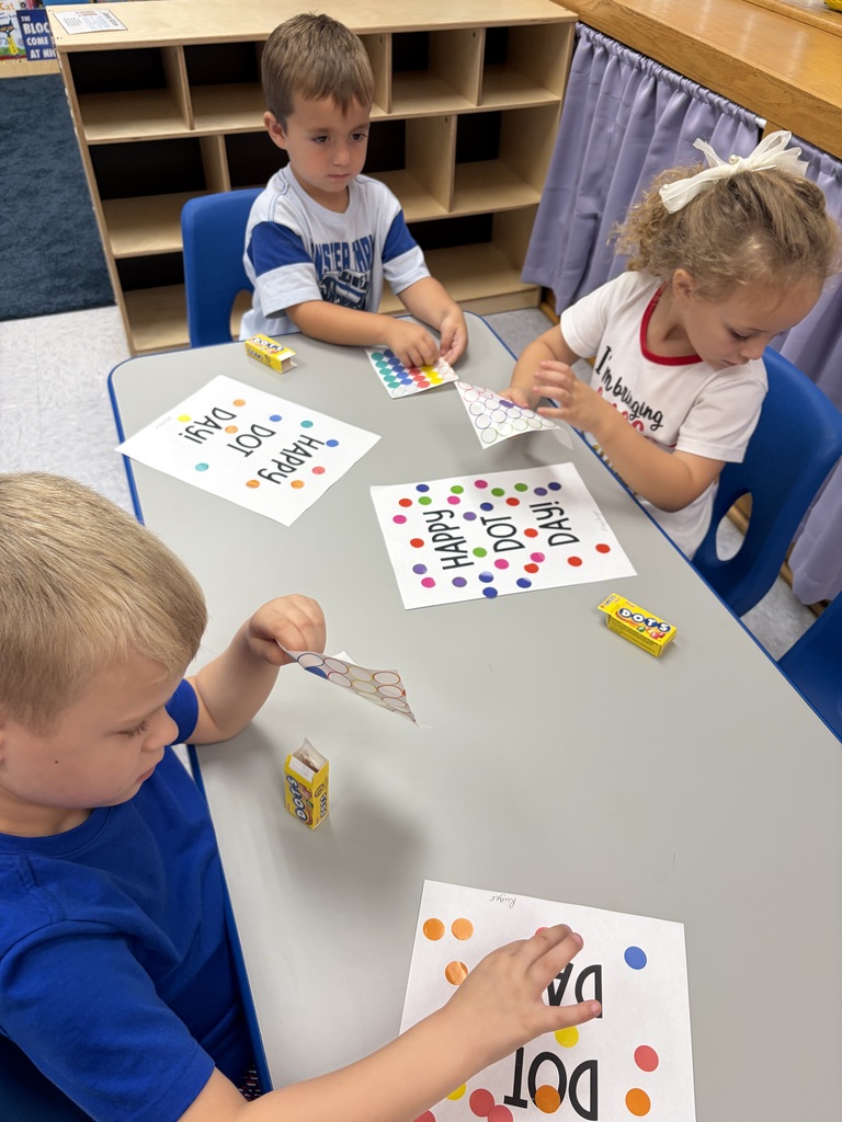 Three children seated at a table with papers, stickers, and other materials. One child holds a paper with the word 'Dog'.