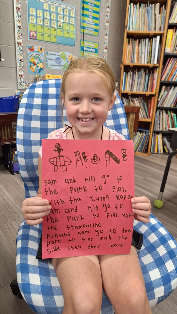 A young girl smiles, holding a pink poster with text. She sits in a blue chair in a classroom with bookshelves in the background.