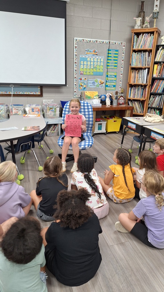 A classroom with children seated in a circle. A girl sits in a chair facing them. Tables and chairs, bookshelves, and a projector screen are in the background.