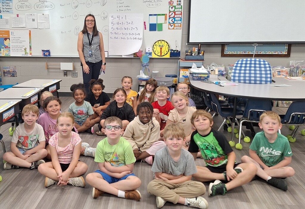 A classroom with children seated in a circle, a teacher standing by a whiteboard, desks, and chairs.