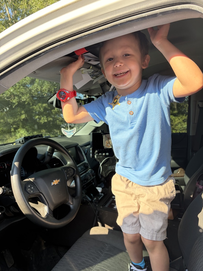 A young boy wearing a blue shirt and khaki shorts is standing inside a vehicle and holding onto the ceiling with a joyful expression.