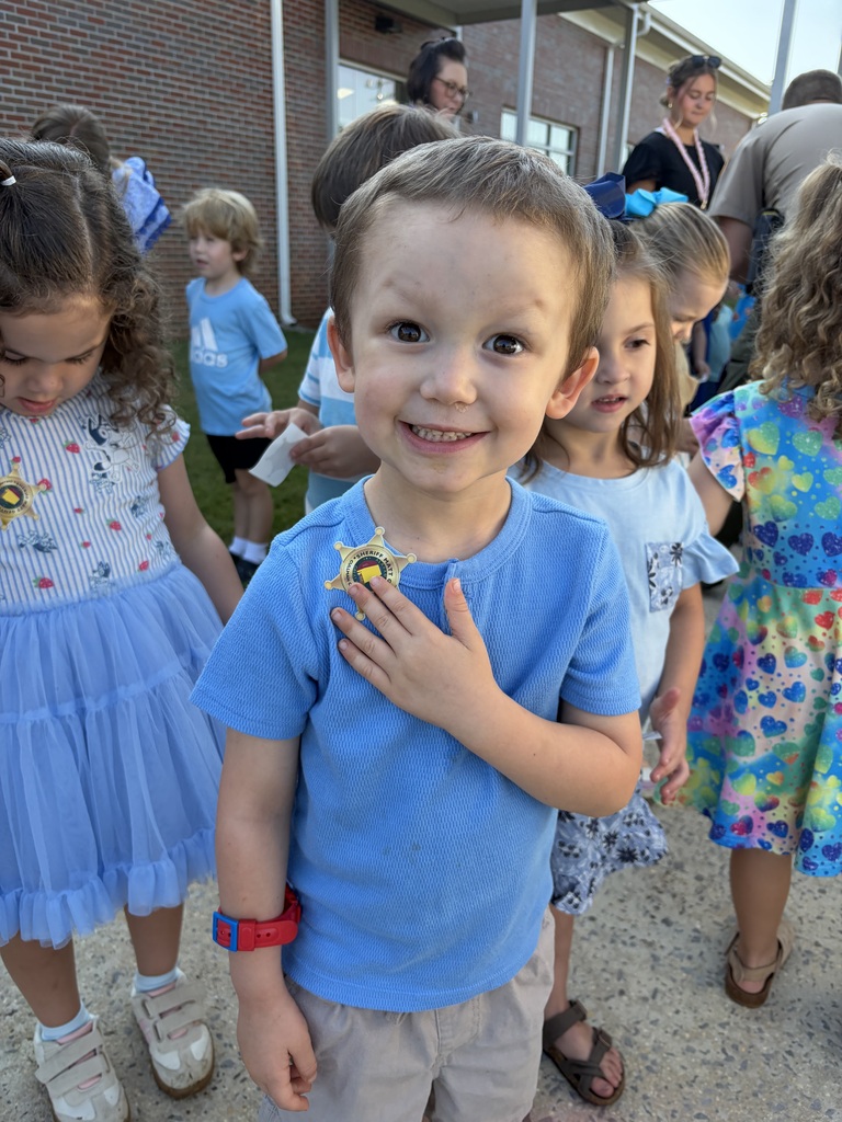 A boy with a badge smiles with children outside a building. Girls in dresses stand behind him.