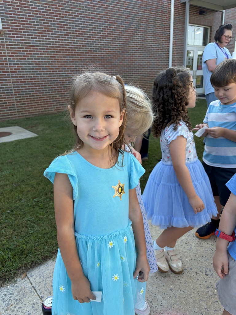 A girl in a blue dress stands in front of a brick building. Other children walk behind her.