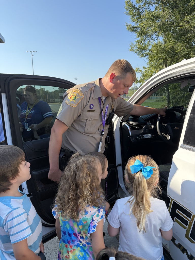 A uniformed police officer shows children the inside of a police car. Trees and a stadium in the background.