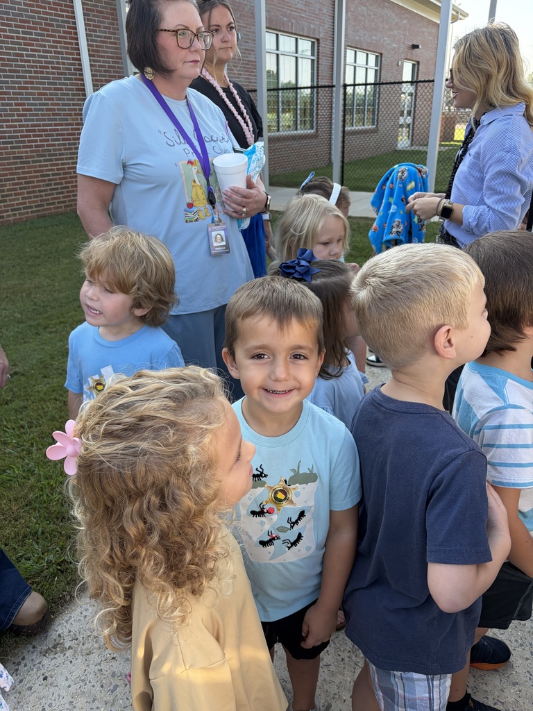 Adults and children stand outdoors near a brick building. A woman holds a cup and ID. Children wear casual clothes.