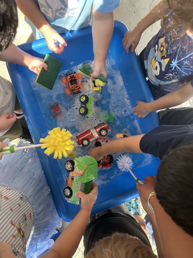 Multiple children engage in a water play activity, using tools to clean toy vehicles in a blue tray.