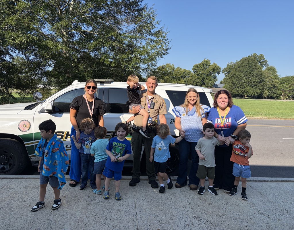 A group of adults and children stand by a police SUV on a sunny day with trees in the background.