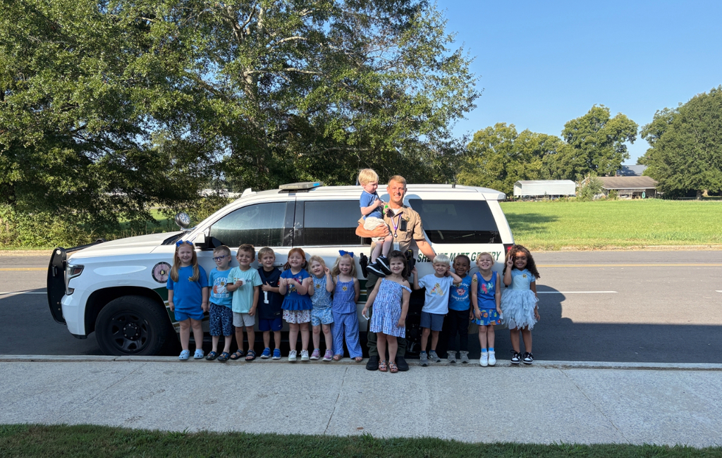 A group of children stands in two rows, smiling. Behind them is a police vehicle. Trees and grass are in the background.