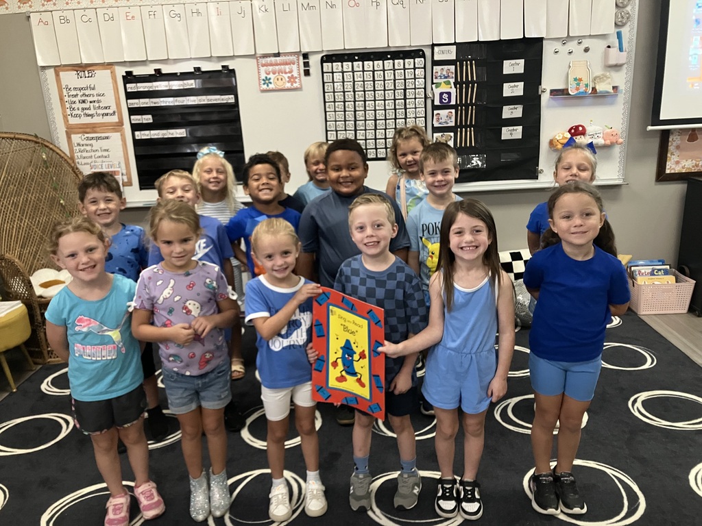 Students in a classroom pose for a photo. They are standing in a line, some holding artwork.