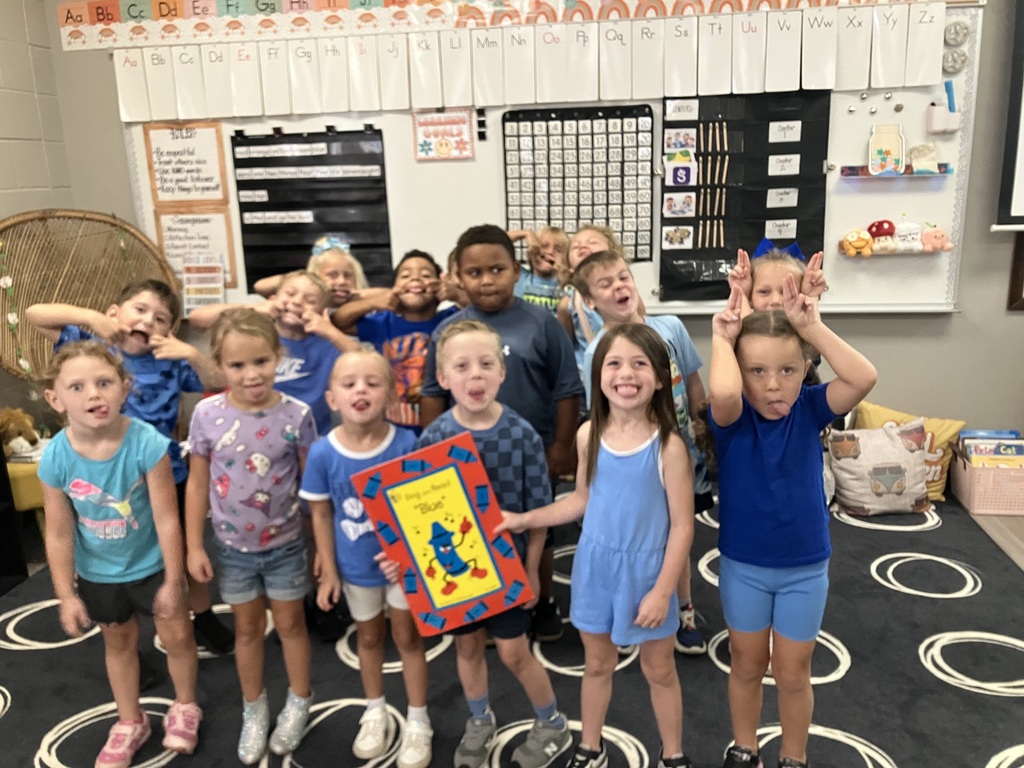 Kids in a classroom, smiling, holding a book, and standing on a black rug with white circles.
