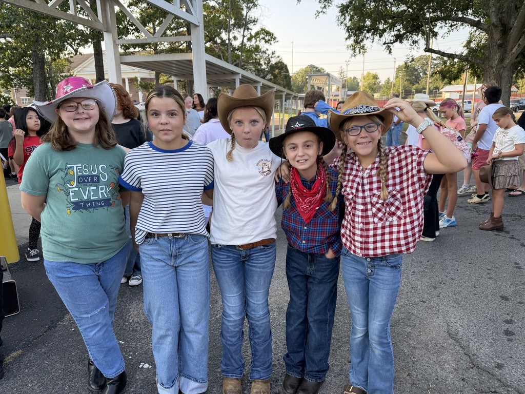 Five girls wearing cowboy hats and boots stand together in a crowd with trees and buildings.