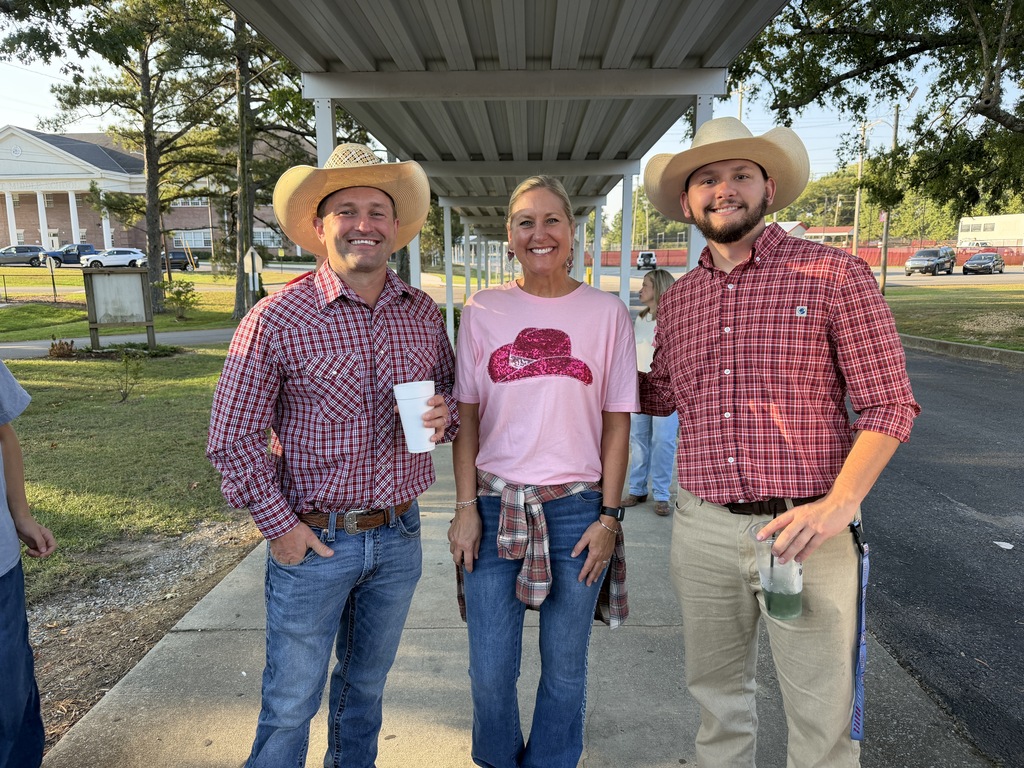 Three people wearing cowboy hats stand outside on a sidewalk, with one holding a cup. A house and cars are visible in the background.