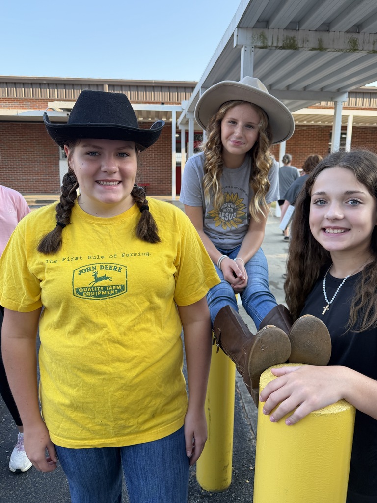 Three girls pose for a picture; one in a yellow shirt, one in a cowboy hat, and another with brown boots on a yellow post.