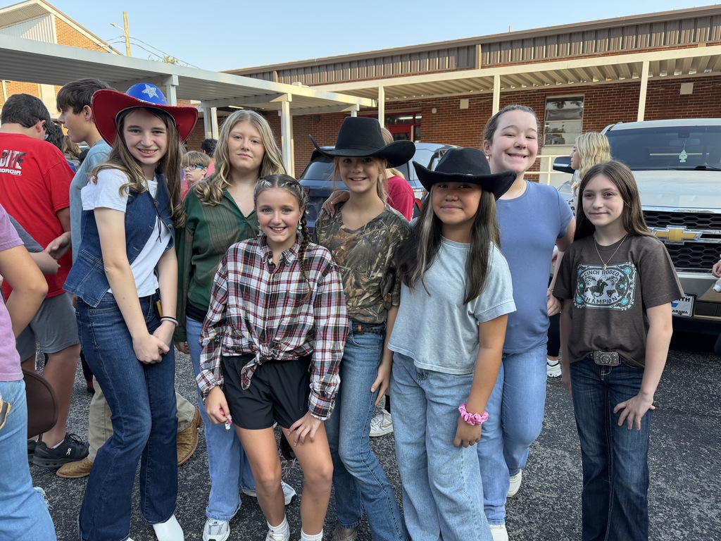 A group of girls stands in a parking lot, wearing hats and jeans. A building is behind them.