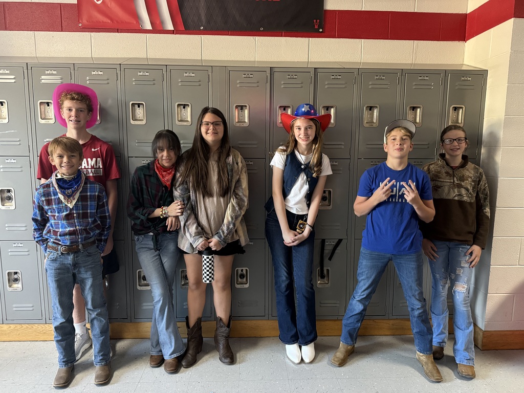 A group of seven people stand in a row in front of lockers, wearing various cowboy hats, boots, and western-themed clothing.