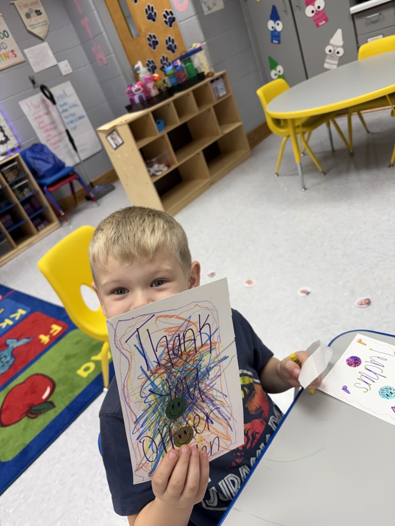 A young boy holding a piece of art sits at a table in a classroom.