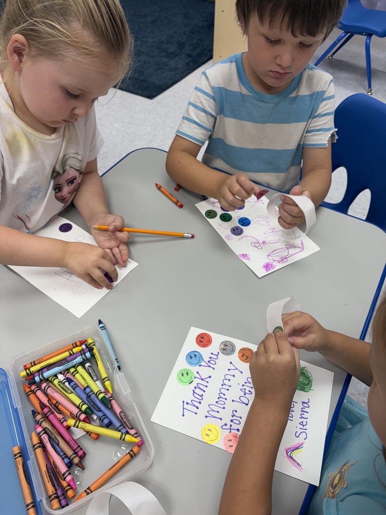 Two children sit at a table, each with a paper and crayons. One child holds a paper with text, while the other works on a paper with a face.