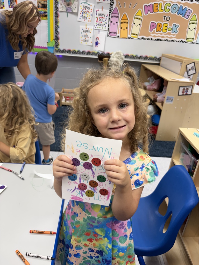 A young girl displays a colorful card in a classroom, with a table of art supplies behind her.