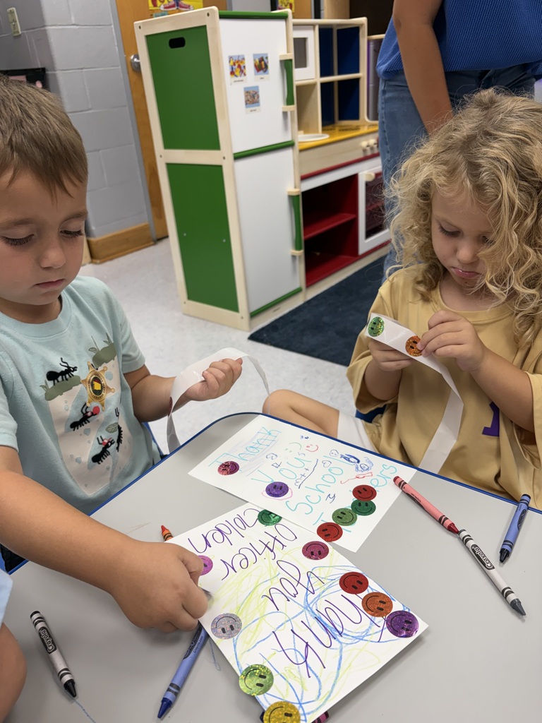 Two young children are seated at a table, using markers to draw and add buttons to a paper.