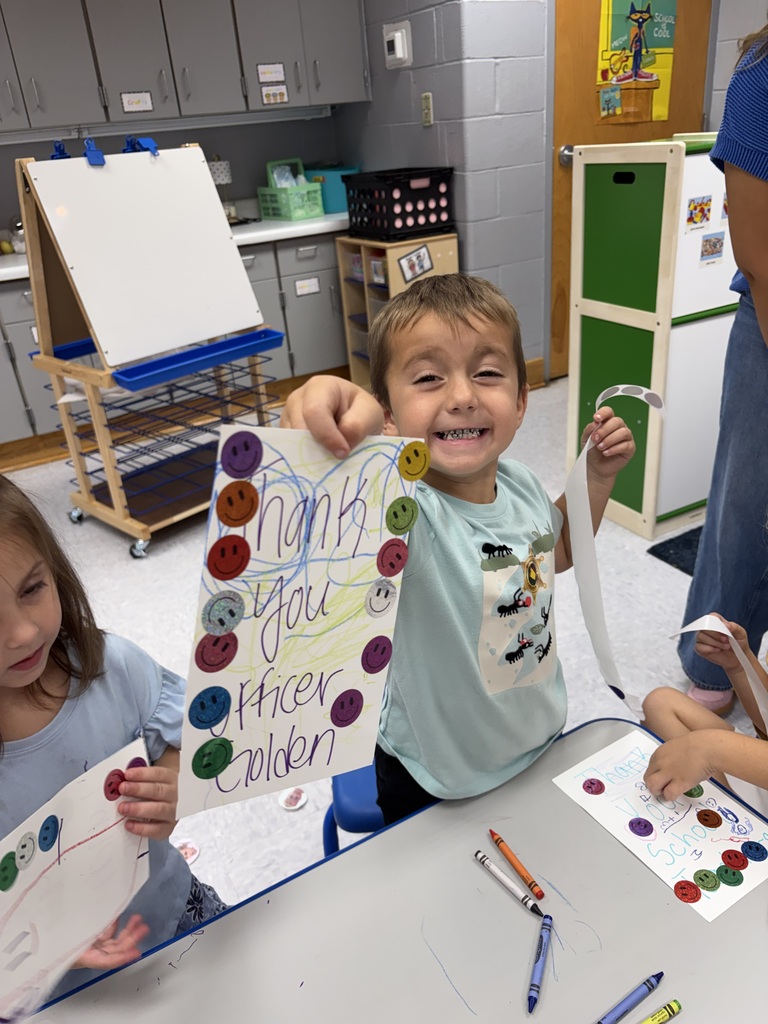 Two children, one holding a paper sign, another holding a crayon, are seated at a table. Behind them, various classroom items are visible.