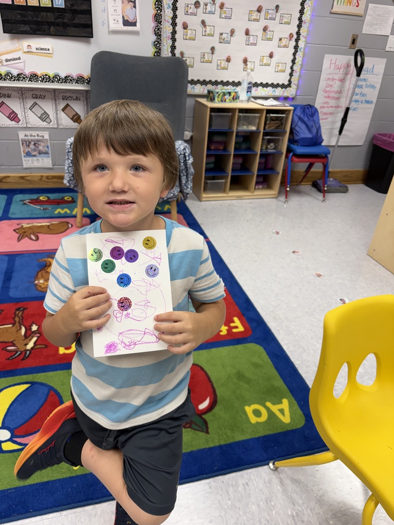 A boy displays a colorful drawing with circles in various colors. He is in a classroom with a rug and chairs.