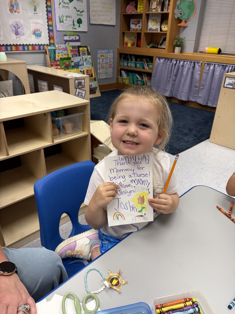 A child holds a paper, smiling. Behind the child is a table and a bookshelf. On the table is a watch, scissors, and crayons.