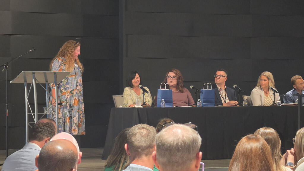 A panel with six people sit at a table on stage, facing an audience. A woman stands on stage with a microphone.