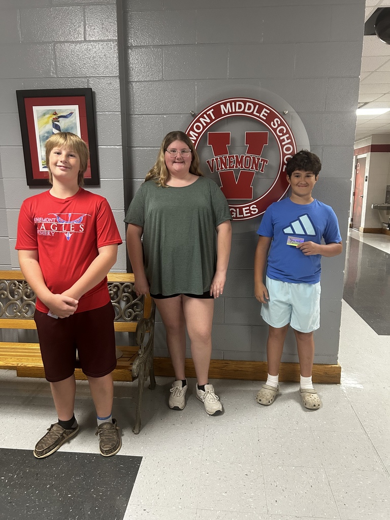 Three individuals stand together in a hallway. The person on the left wears a red shirt and shorts, the middle person wears a green shirt and shorts, and the person on the right wears a blue shirt and shorts.