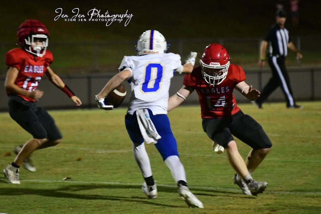 Three football players on a field. One holds a ball; another tackles. A referee walks in the background.