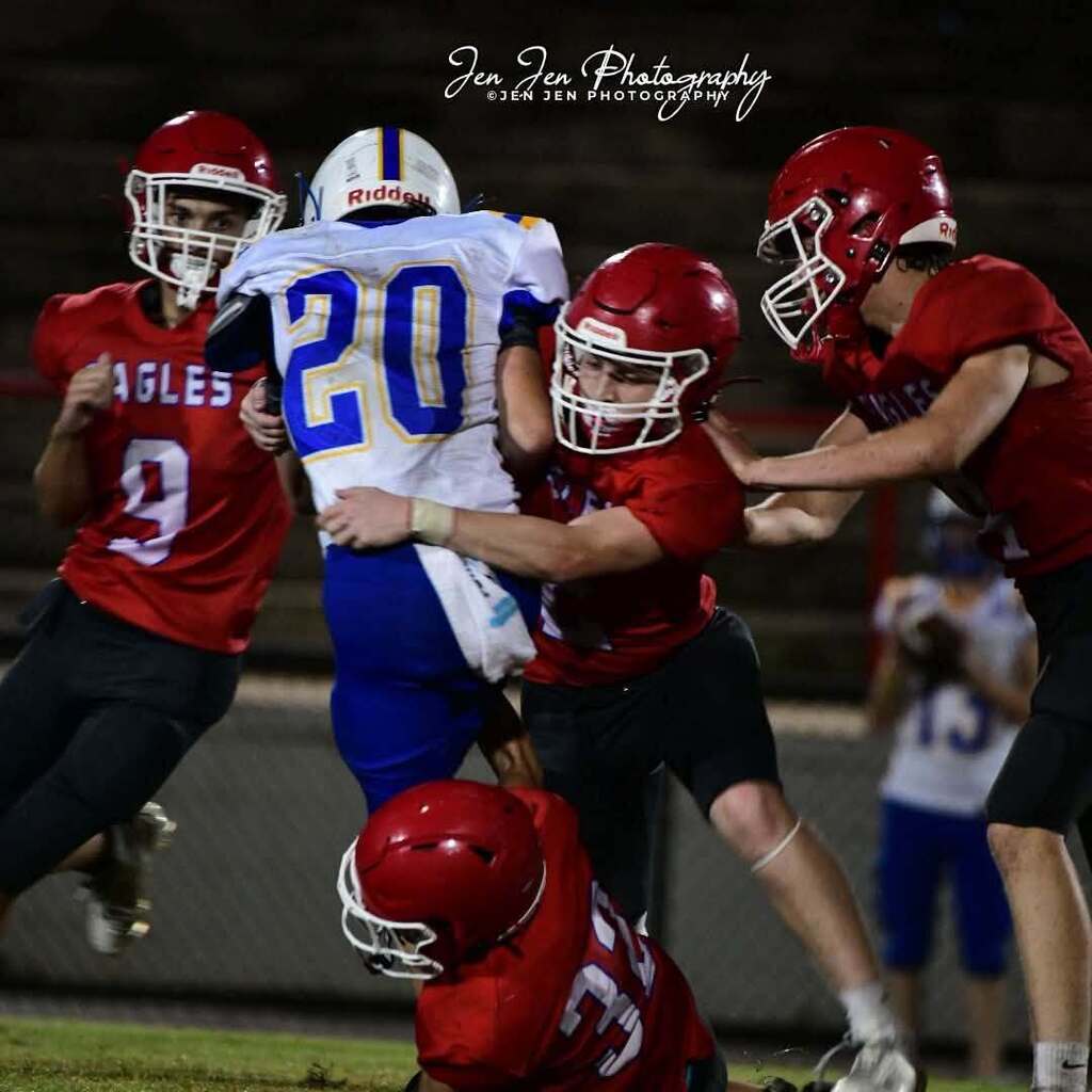 Four football players, two wearing red jerseys, two wearing blue jerseys. They are in mid-action, tackling, and falling.