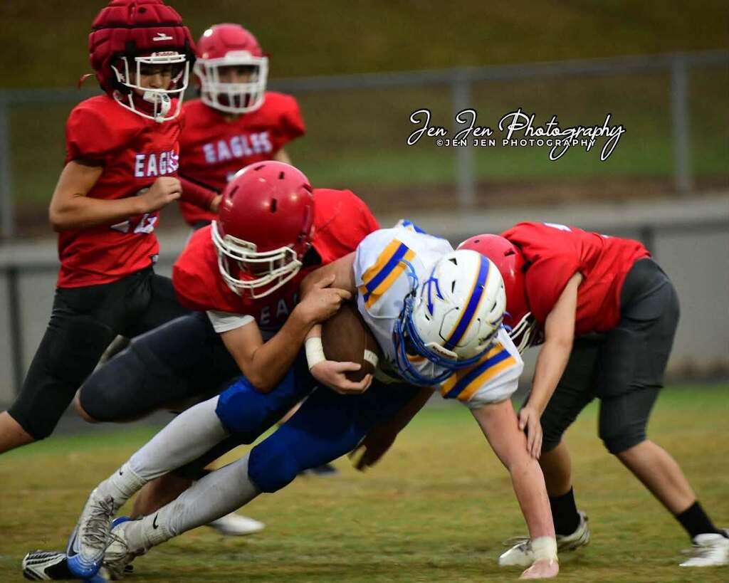 Several youth football players in red and blue uniforms collide on a field. The player in red is tackling the player in blue.
