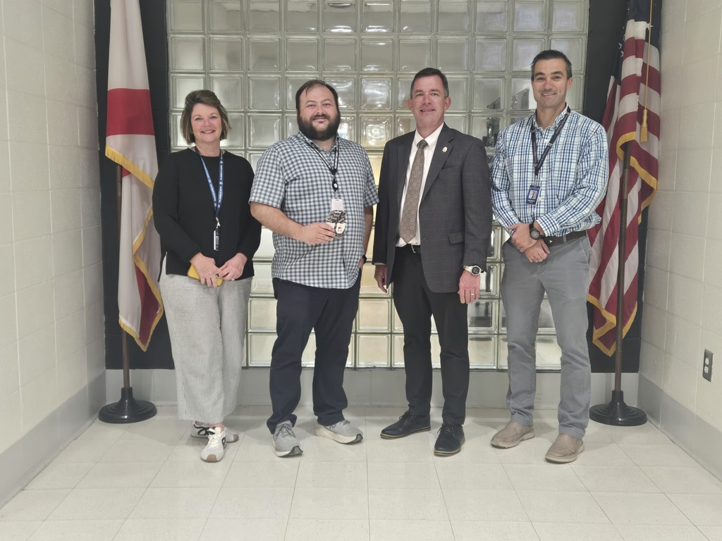 Four people stand in a hallway. Two flags are behind them. The floor is white, and the walls are glass.