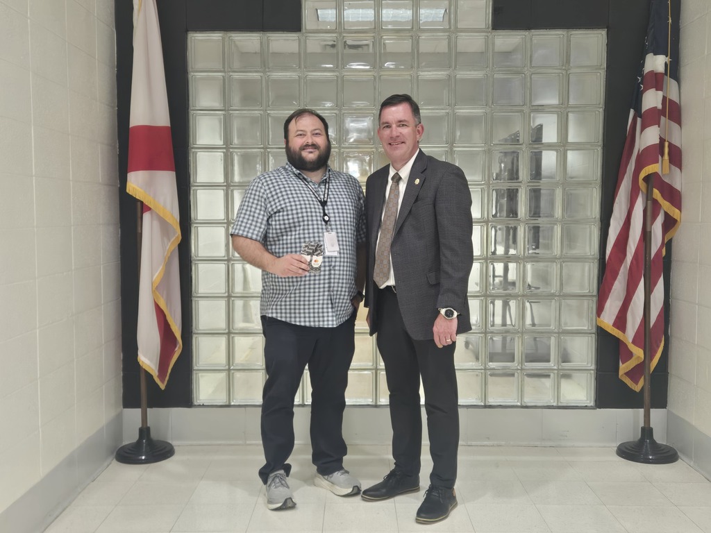 Two men stand in a room with white tile flooring and two flags behind them. One wears a suit and tie, the other a casual shirt and jeans.