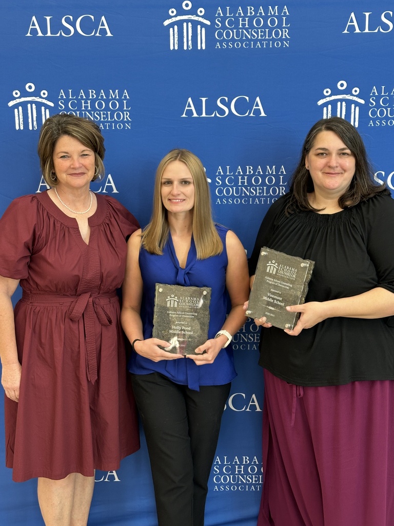 Three women stand behind a blue backdrop with text. Each holds a plaque. They smile and look at the camera.