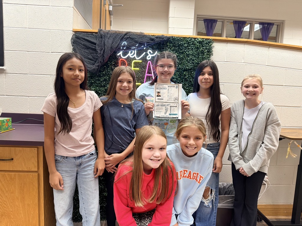 A group of young girls smiling for a photo in a classroom. One holds a certificate. Behind them is a decorated wall.