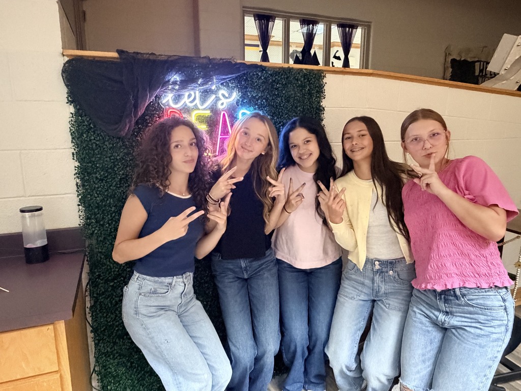 Five women pose with peace signs in front of a green wall with a neon sign.