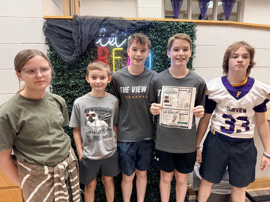 Five kids stand in front of a green wall. Two boys wear sports jerseys. Three boys wear shirts with printed designs.