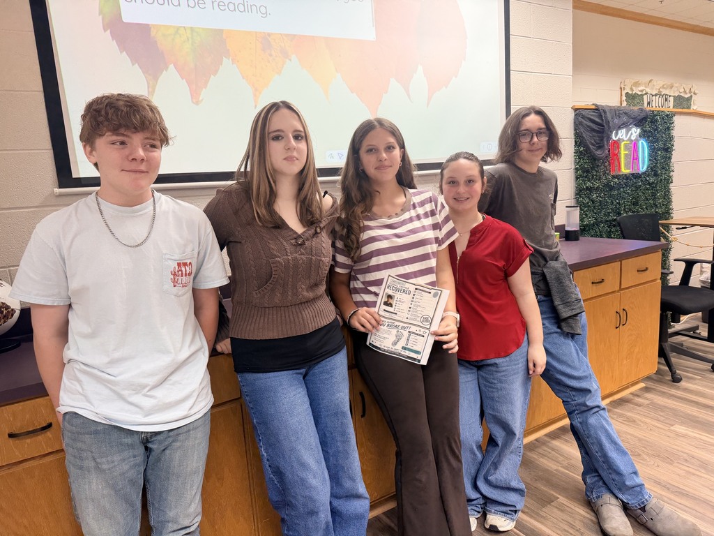 Five students standing behind a counter in a classroom with a projector screen showing text.