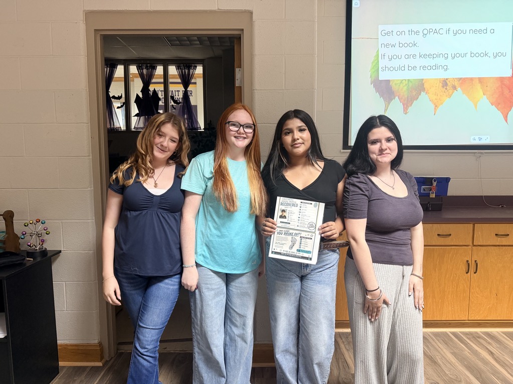Four women stand together in front of a projector screen and cabinets, holding a paper. They wear casual clothes and glasses.