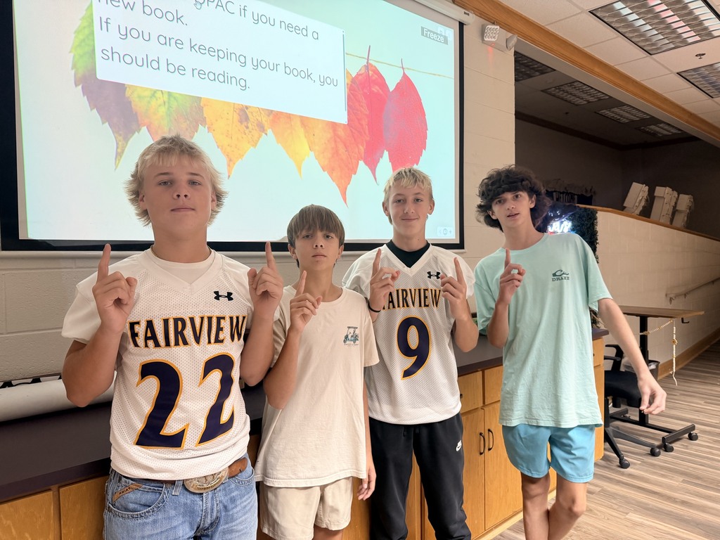 Four boys stand in a classroom, posing for a photo. One holds a peace sign, others hold numbers 22, 9, and 20. Behind them is a projector screen.