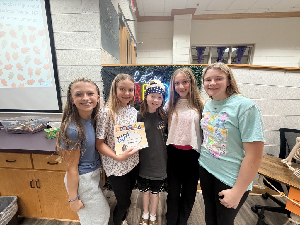 Five girls stand together in a room with a presentation screen. One girl holds a paper with writing.