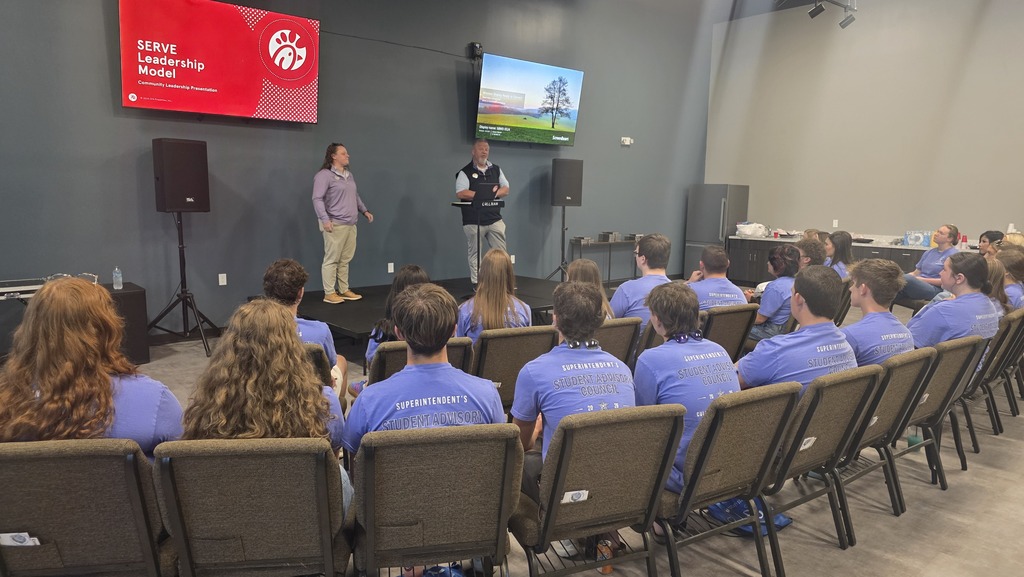 A classroom with students seated in rows. Two people stand on a stage with a screen and speakers behind them.