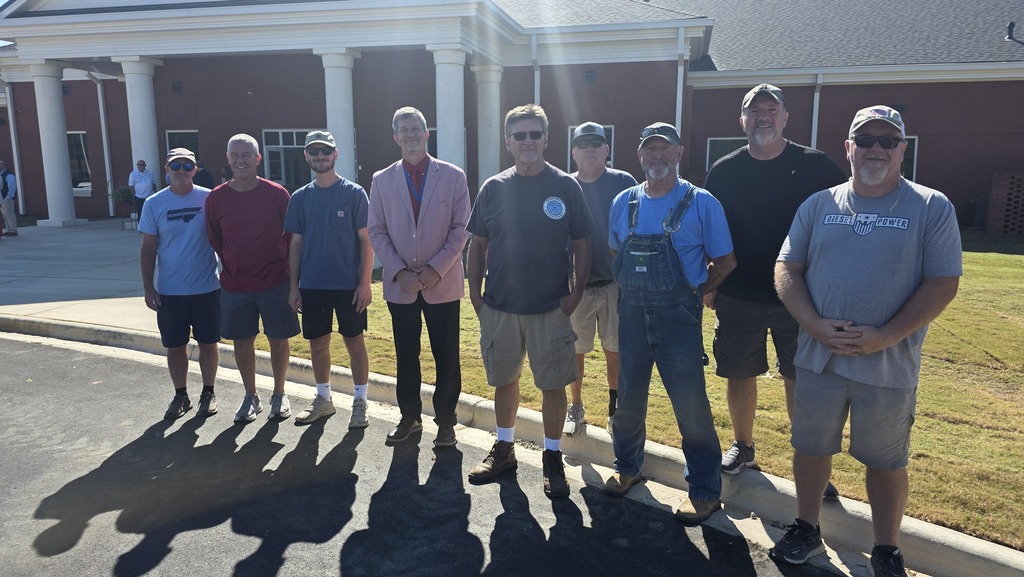 A group of men in casual attire stands outside a building with a large lawn in the background.