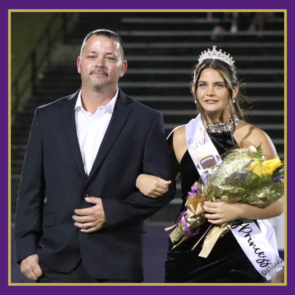 A man in a black suit and a woman in a black dress and tiara pose together. The woman holds a bouquet.