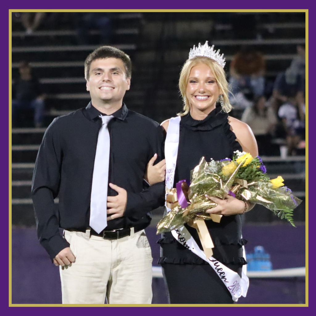 Man and woman in black attire and crown stand together, holding a bouquet of flowers in a stadium.
